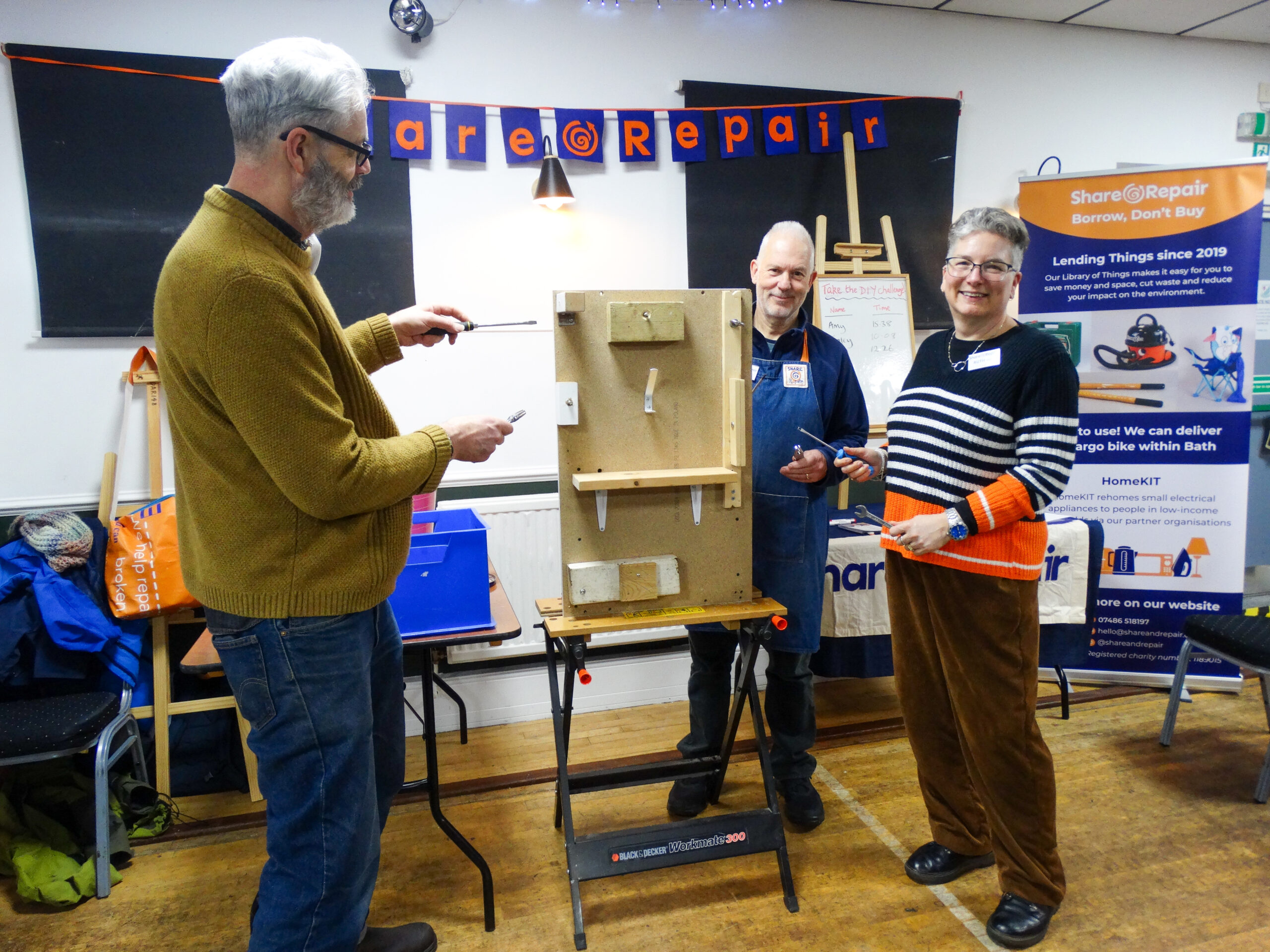 Julian, Dick and Ruth standing around a DIY challenge which is a wooden box with shelves and blocks of wood attached to it, at the Youth Guarantee Roadshow